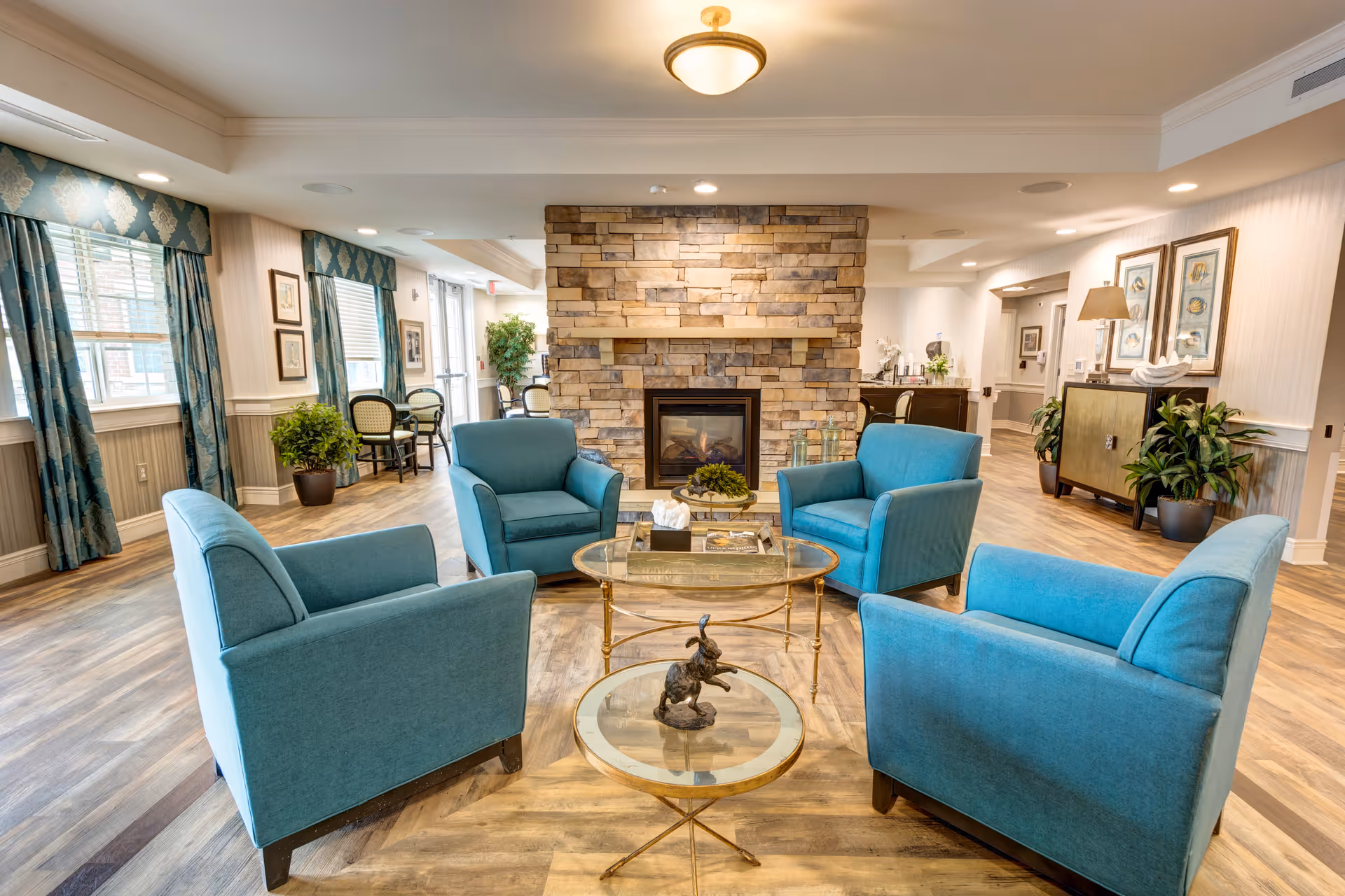 A cozy living room area with four blue armchairs arranged around two glass-top coffee tables. The room features a stone fireplace in the center background, wood flooring, large windows with blue patterned curtains, and various plants and framed artwork on the walls.