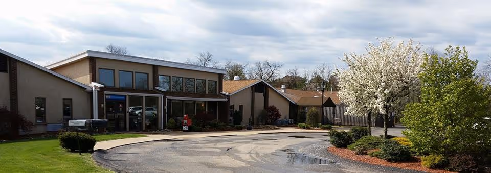 Exterior view of a single-story building with large windows and a sloped roof, surrounded by a paved driveway and landscaped greenery including bushes and a tree with white blossoms under a cloudy sky.
