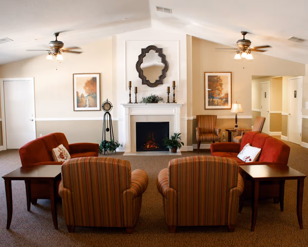 A cozy living room area in Bradford Place featuring two red sofas, two striped armchairs, a white fireplace with a decorative mirror and candles above it, two framed landscape paintings on the walls, a standing clock, and ceiling fans with lights.