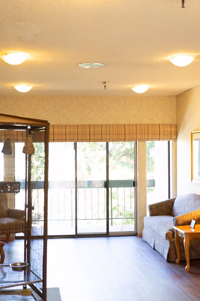 A bright living room area with a large sliding glass door leading to a balcony. The room features a patterned sofa with a wooden side table, and a birdcage is partially visible on the left side. The ceiling has multiple round light fixtures, and the window has a striped valance.