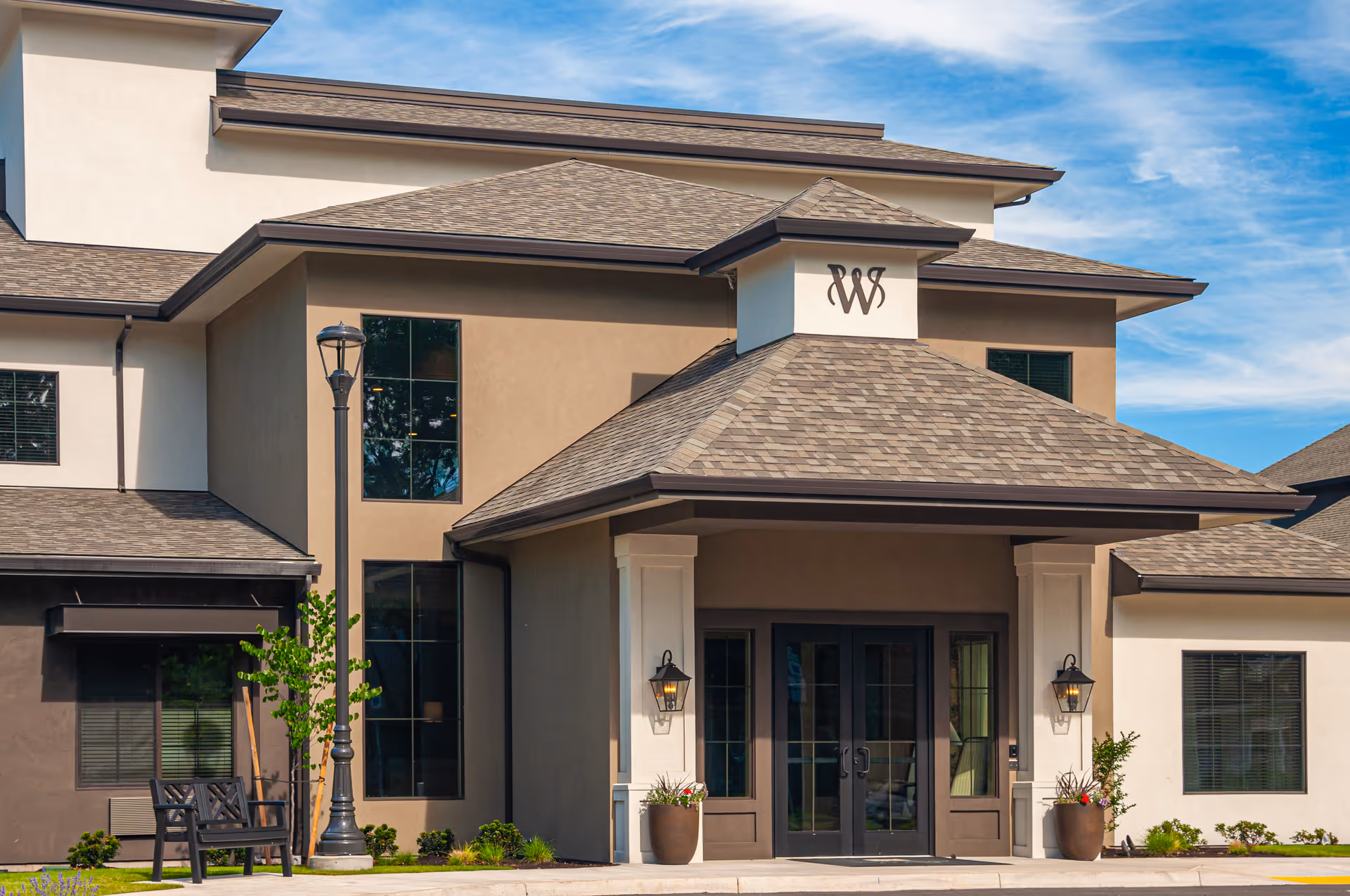 Exterior view of Weatherly Inn Independent Senior Living building entrance with a covered porch, double glass doors, two wall-mounted lanterns, potted plants, a bench, and a street lamp under a blue sky with some clouds.