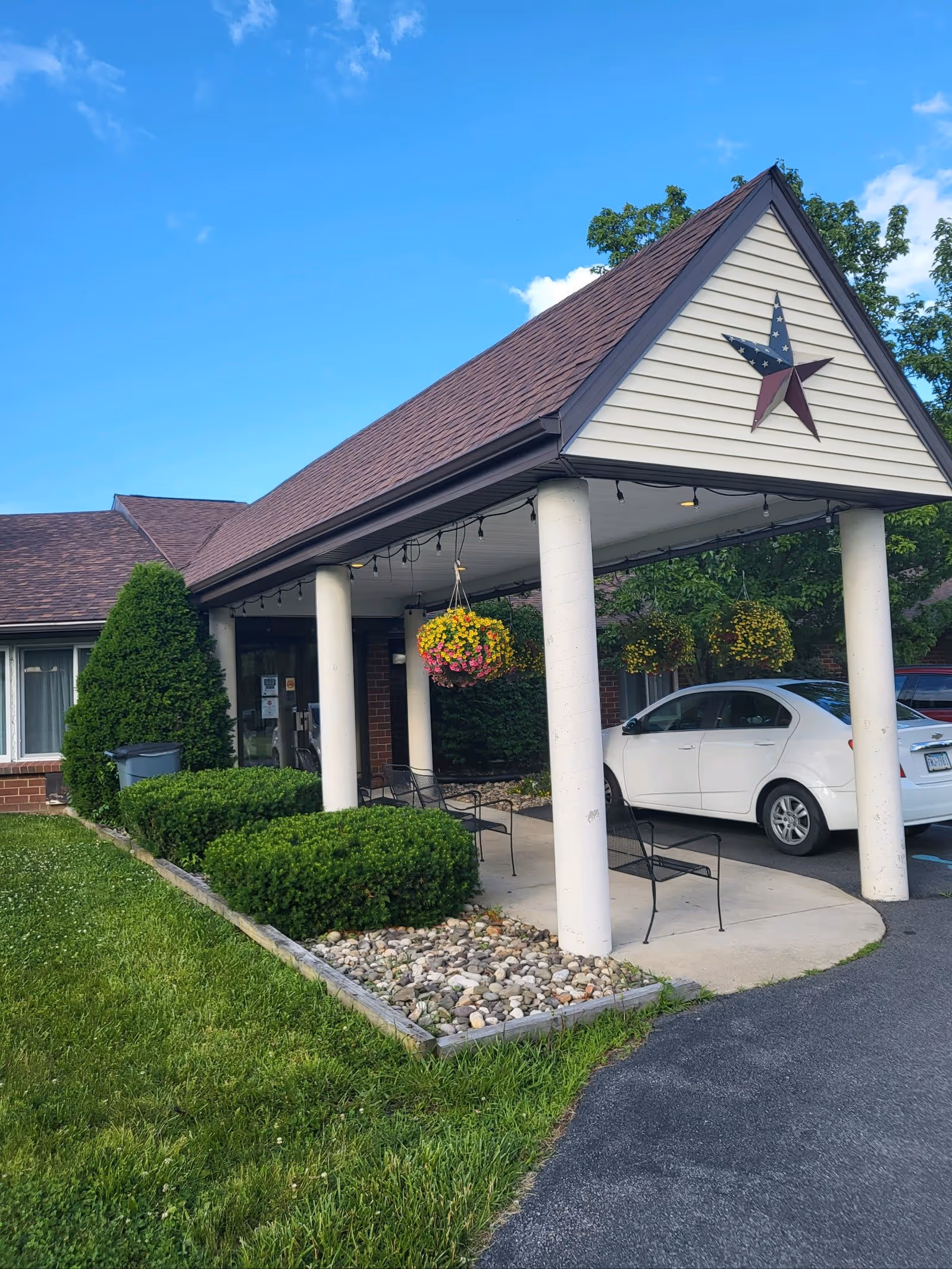 Entrance of a building with a covered porch supported by white columns, hanging flower baskets, and two black metal chairs. A white car is parked under the porch. The building has a brown shingled roof and a decorative star on the front gable. Green bushes and a small rock garden border the walkway leading to the entrance.