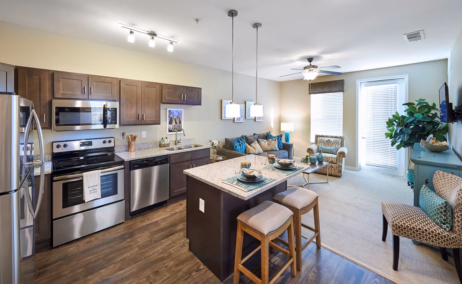 Open-concept living space featuring a modern kitchen with stainless steel appliances, dark wood cabinets, and a granite island with two cushioned stools. The living area includes a sofa with blue and beige pillows, an armchair, a coffee table, a ceiling fan, and a TV mounted on the wall. Large windows and a glass door with blinds allow natural light into the room.