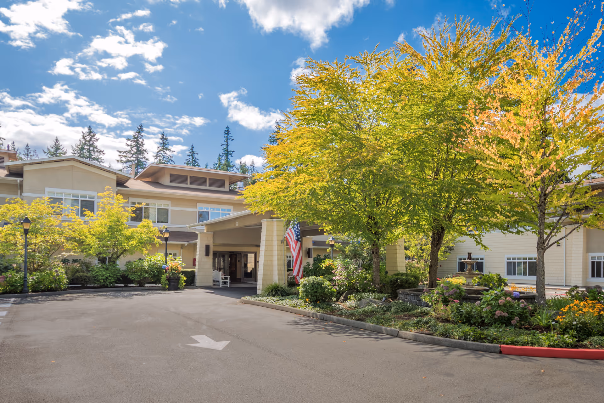 Exterior view of Sunrise of Redmond senior living facility entrance with a covered drop-off area, surrounded by trees and landscaped greenery under a partly cloudy blue sky.
