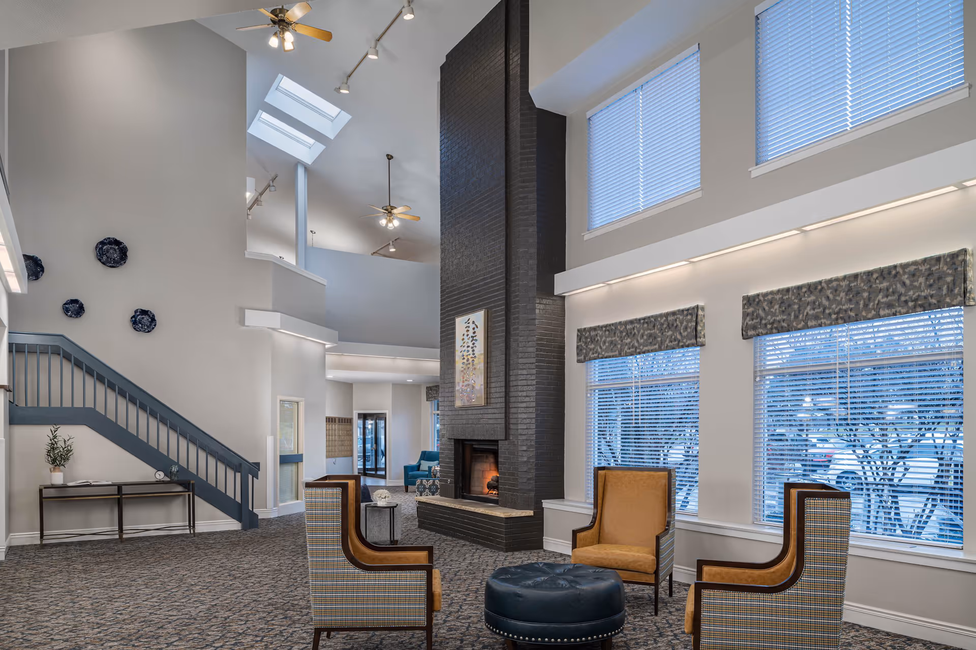 A spacious senior living facility common area with high ceilings, large windows with blinds, and a tall black fireplace in the center. The room features a seating arrangement with four armchairs around a round ottoman. There is a staircase with a blue railing on the left side and decorative plates on the wall above a small console table with a plant and clock.