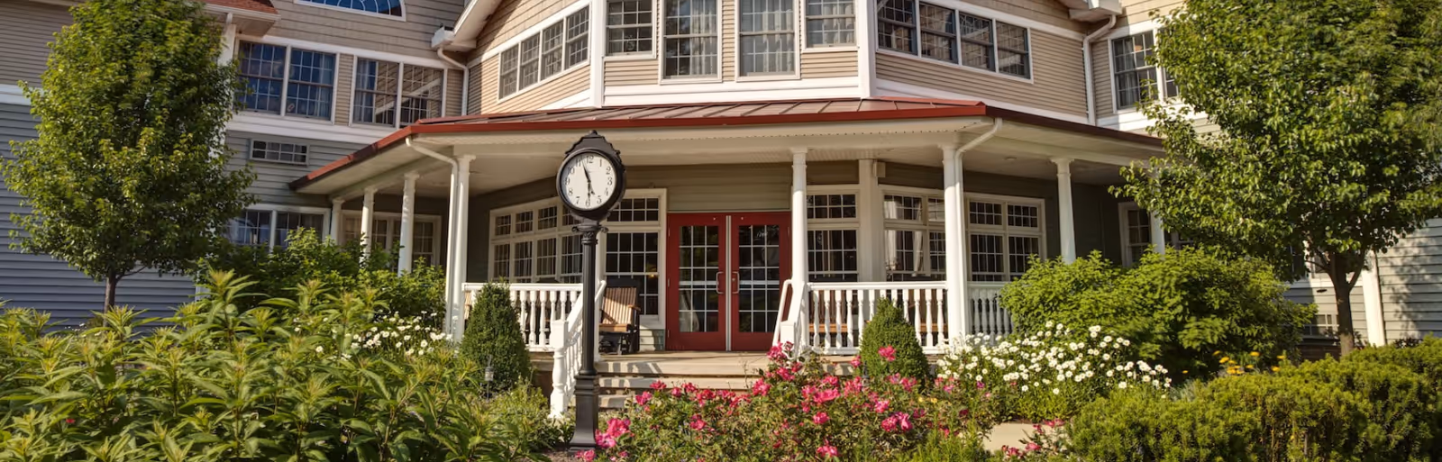 Front exterior view of Briar Hill Health Care Residence showing a large porch with white railings and red double doors. The building has multiple windows and a red roof over the porch. There is a black street clock in front of the porch surrounded by well-maintained greenery and colorful flowers.