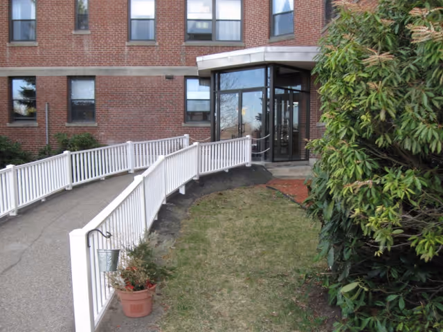 Entrance to a brick building with a glass door and a white ramp with railings leading up to it. There is a small potted plant on the left side of the ramp and green bushes on the right side of the image.