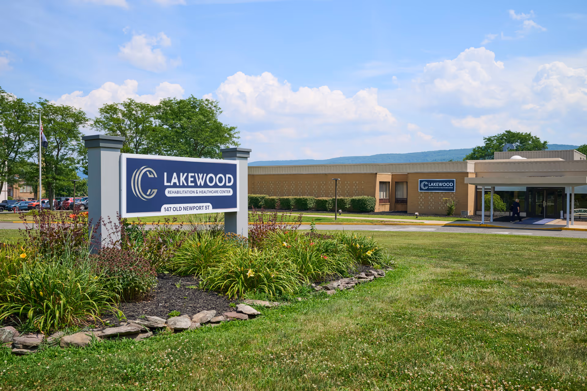 Exterior view of Lakewood Rehabilitation and Healthcare Center with a large sign in the foreground surrounded by landscaping and the building in the background under a partly cloudy sky.