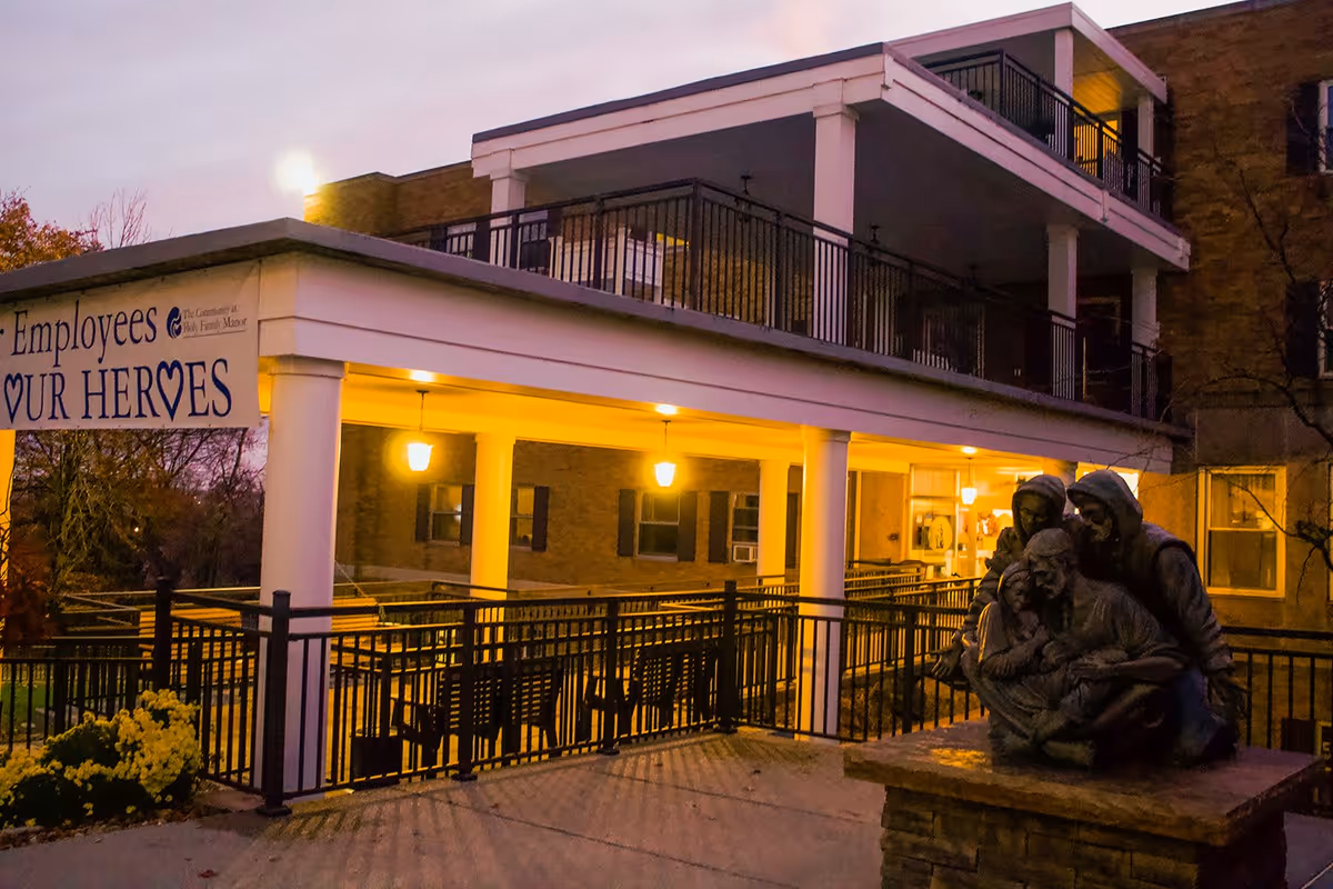 Evening view of the exterior of The Community at Holy Family Manor building with a covered patio area illuminated by warm lights. A statue of a group of people is visible in the foreground on a stone pedestal. A banner on the building reads 'Employees Our Heroes' with a heart symbol.