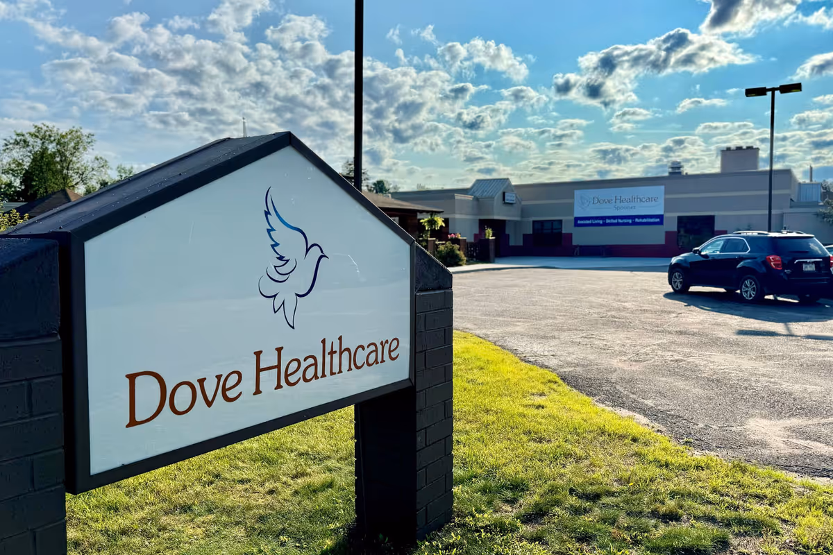 Outdoor view of Dove Healthcare - Spooner facility with a large sign in the foreground featuring a dove logo and the text 'Dove Healthcare'. The building and parking lot are visible in the background under a partly cloudy sky.