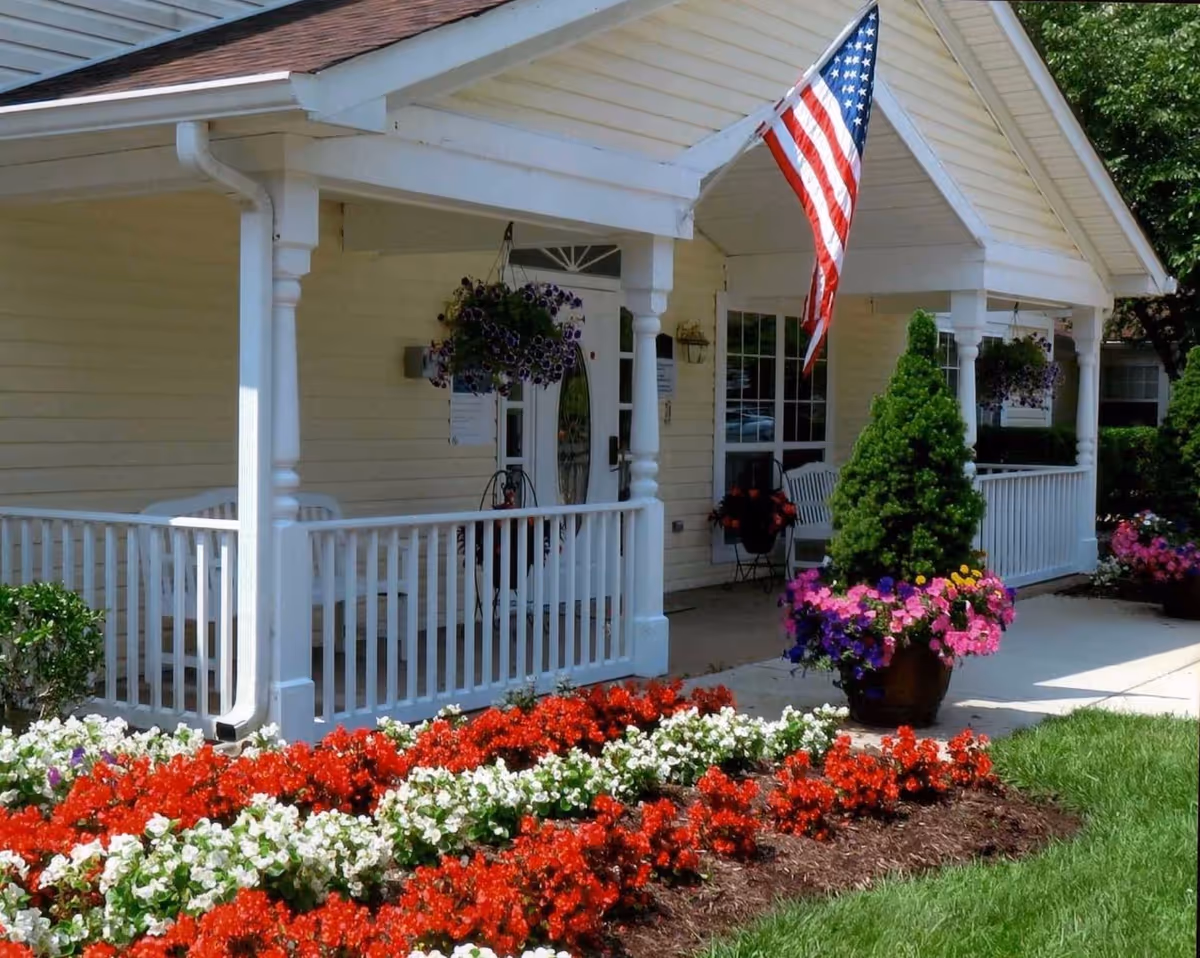 Front porch of a yellow building with white railings and columns, decorated with hanging flower baskets and a large American flag. There are vibrant red and white flowers planted in the garden bed in front of the porch, along with a potted plant with colorful flowers near the entrance.