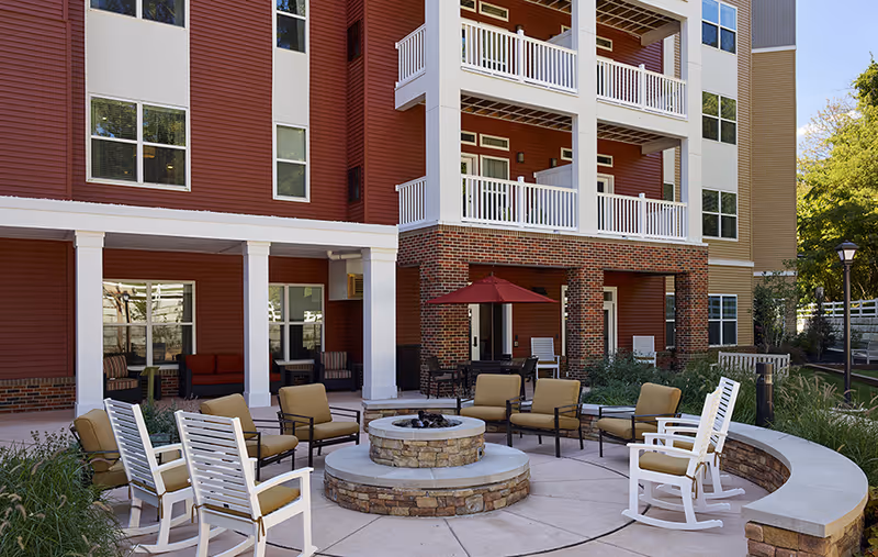 Outdoor seating area at a senior living facility with cushioned chairs and rocking chairs arranged around a circular stone fire pit. The background shows a multi-story building with balconies, red and beige siding, and large windows. There is also a red patio umbrella and some greenery around the seating area.