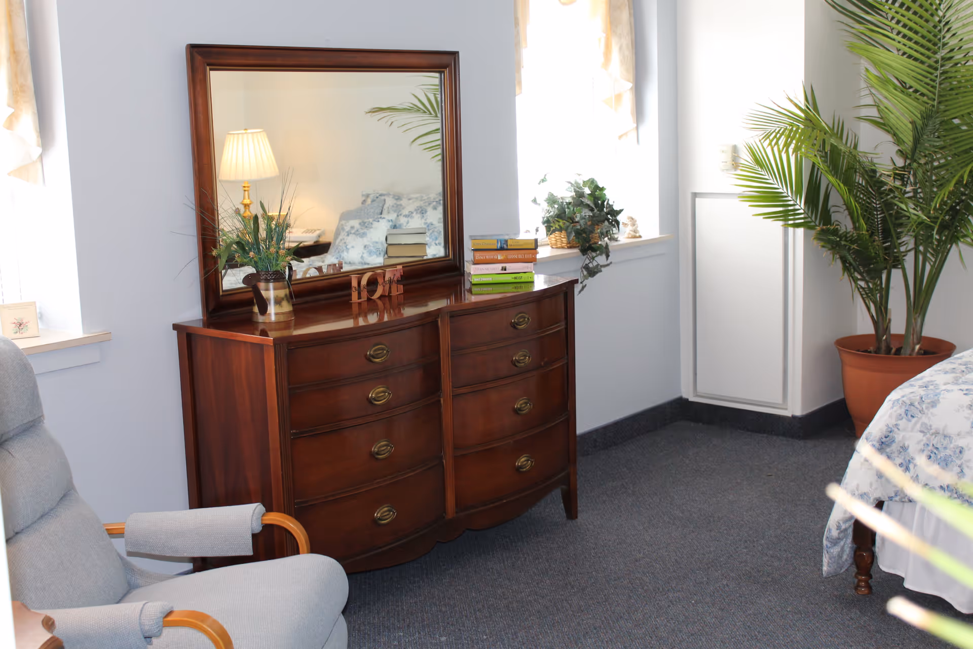A cozy bedroom corner featuring a wooden dresser with a large mirror, a stack of books, a decorative plant, and a small 'LOVE' sign. To the left is a comfortable gray armchair, and to the right, part of a bed with floral bedding and a large potted plant. The room has light-colored walls and carpeted flooring.