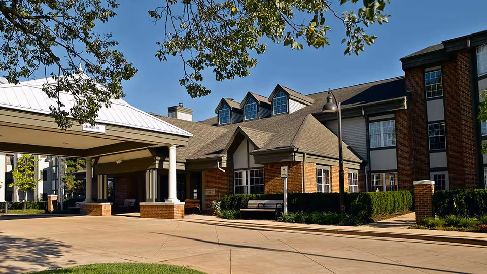 Exterior view of Atria Kinghaven senior living facility showing a covered entrance with white pillars, brick and siding walls, multiple windows, and a clear blue sky with tree branches overhead.