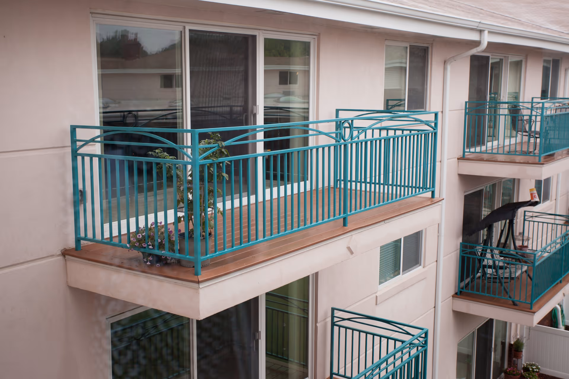 Exterior view of a senior living facility showing multiple balconies with teal railings attached to beige walls. Each balcony has sliding glass doors leading inside, and some balconies have potted plants and outdoor furniture.