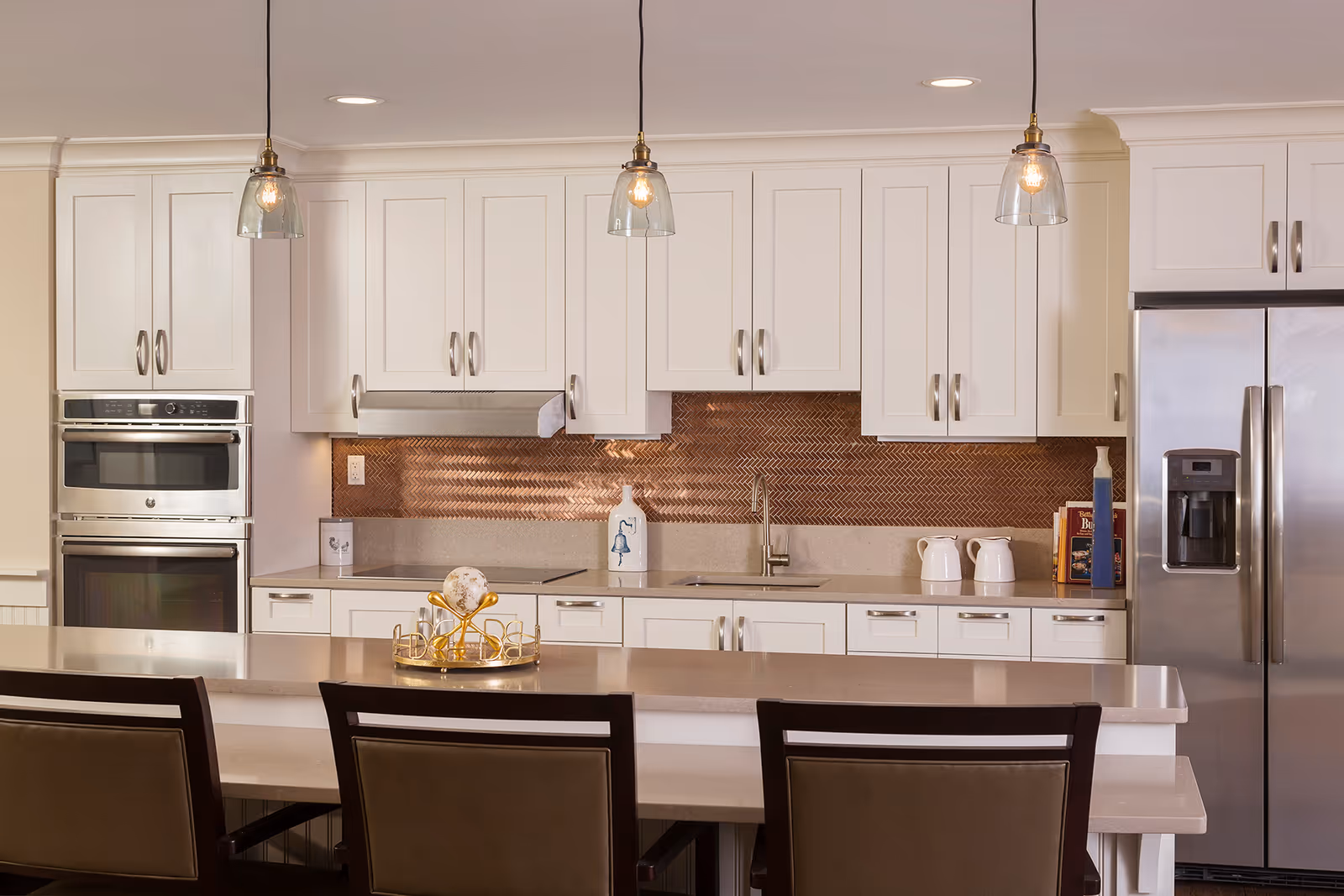 Modern kitchen with white cabinetry, stainless steel double oven and refrigerator, a copper-colored backsplash, three pendant lights hanging over a countertop with three chairs, and various kitchen items on the counter.