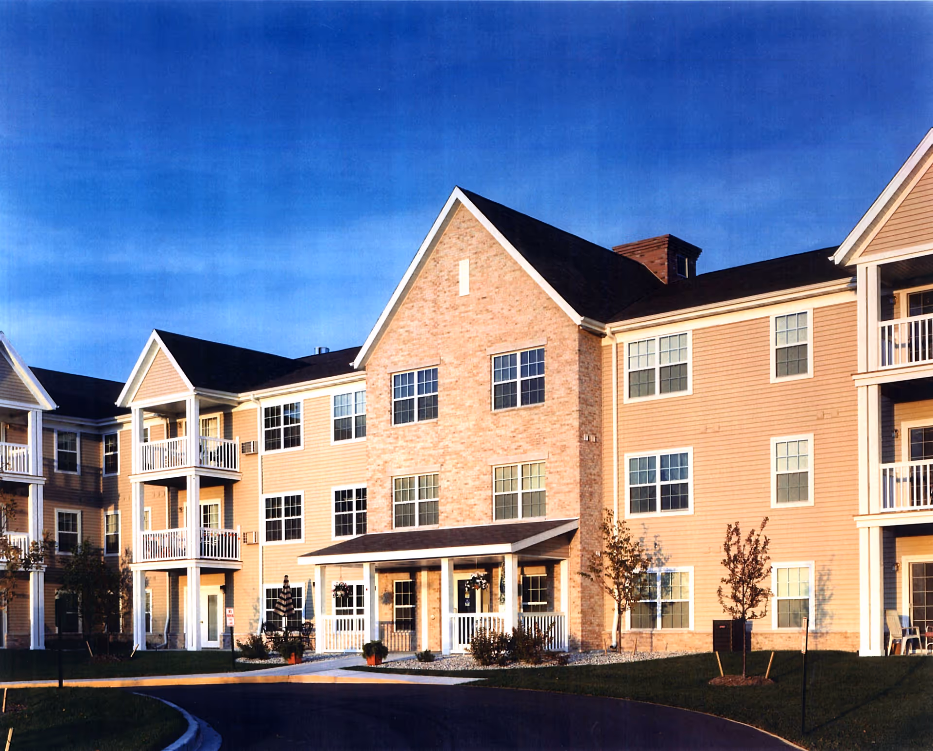 Exterior view of a multi-story senior living facility building with beige siding and brick accents, featuring multiple windows and balconies, a covered entrance, and a curved driveway in front under a clear blue sky.