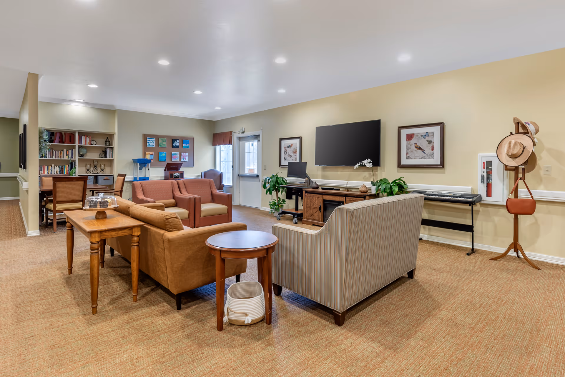 A cozy living room area in a senior living facility with multiple armchairs and sofas arranged around a wooden coffee table and side table. The room features a large flat-screen TV mounted on a beige wall, framed bird artwork, a keyboard on a stand, and a coat rack with hats and a purse. There is a bookshelf and a table with chairs in the background, along with a door and windows letting in natural light.