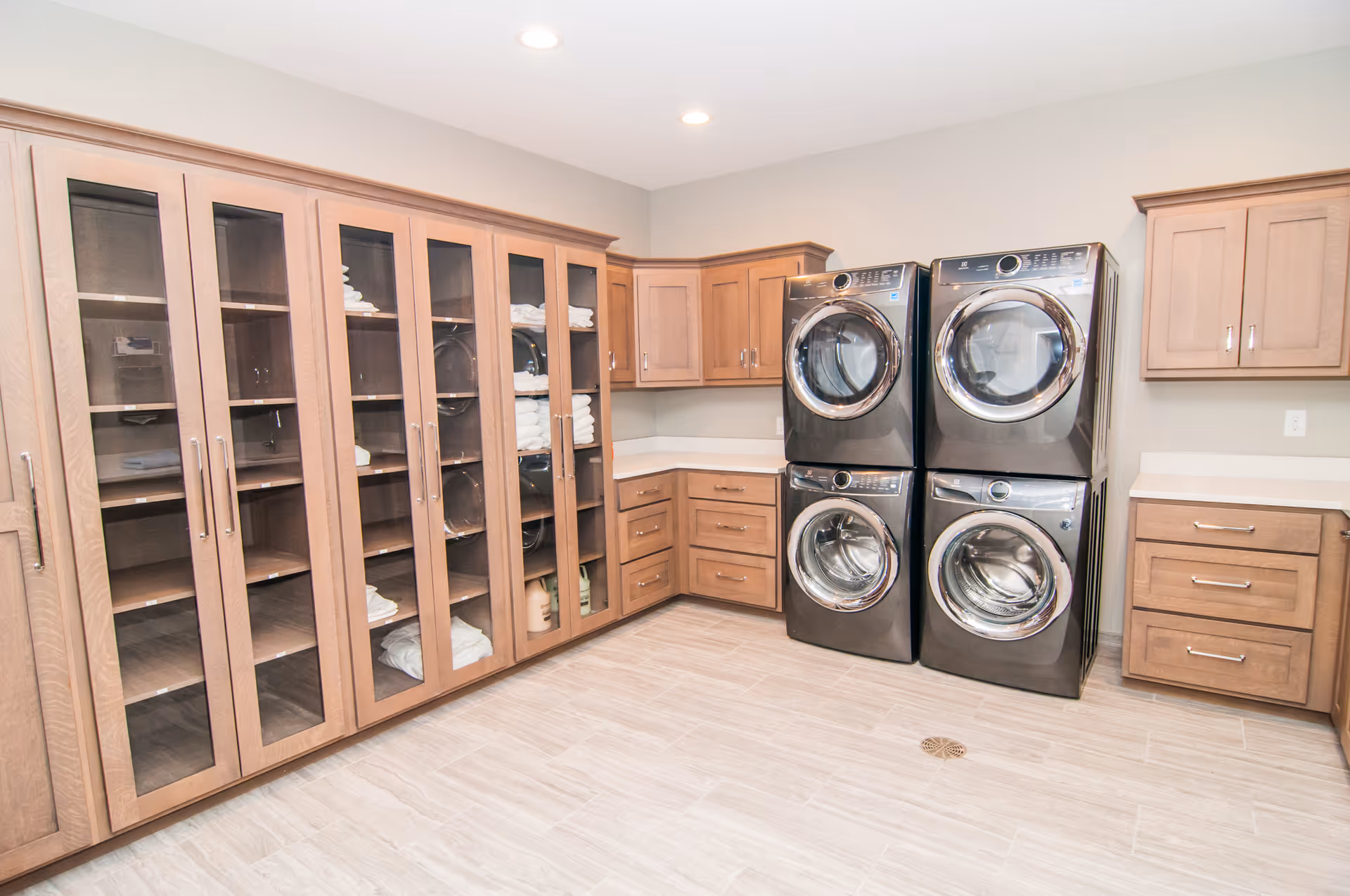 A spacious laundry room with four modern front-loading washers and dryers stacked in pairs. The room features light wood cabinetry with glass doors displaying neatly folded towels and linens. The floor is tiled in a light neutral color, and the walls are painted a soft beige. Recessed ceiling lights illuminate the clean and organized space.