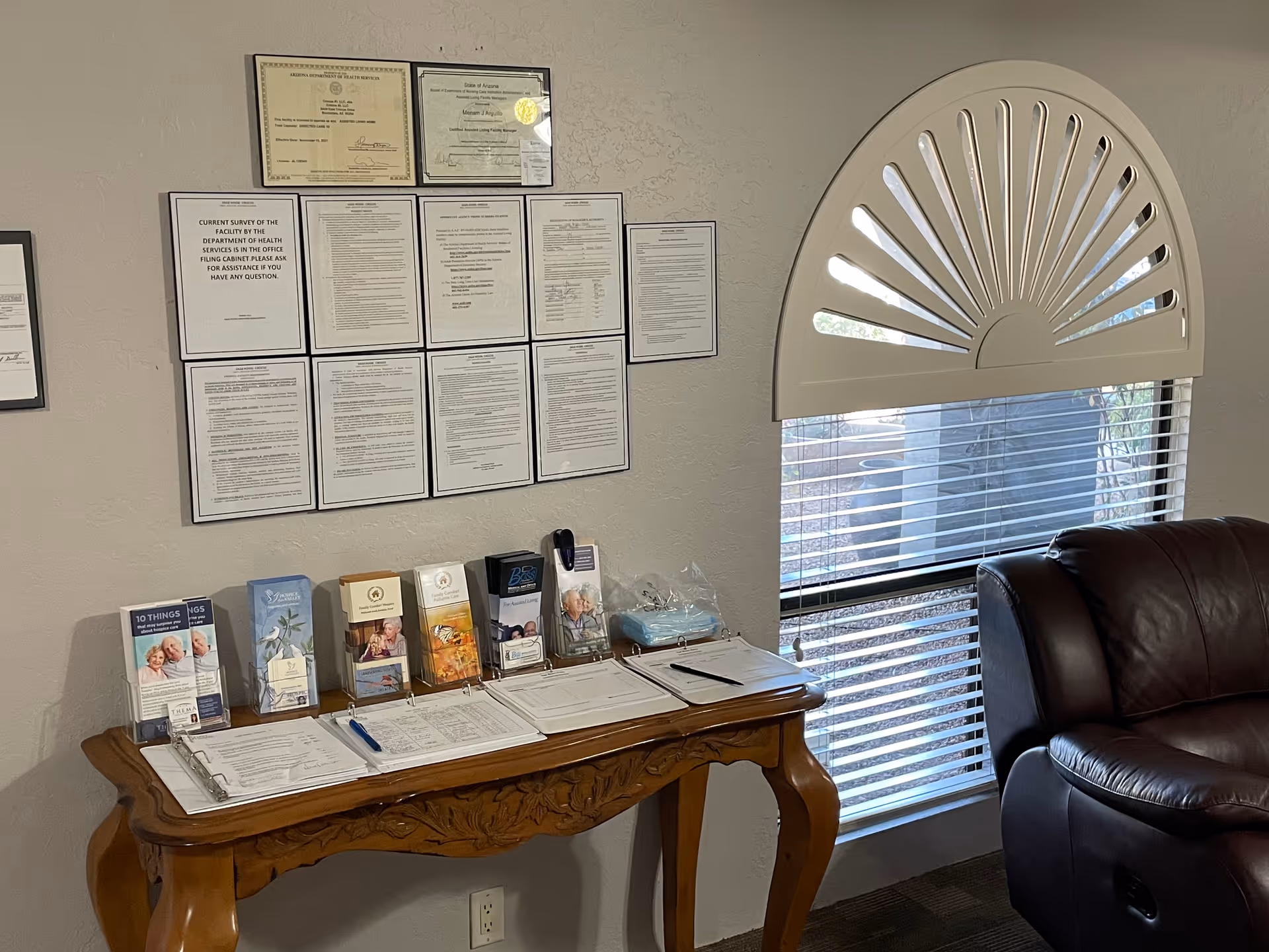A wooden table against a wall with multiple informational brochures and clipboards with papers and pens. Above the table, several framed certificates and documents are displayed on the wall. To the right, there is a window with blinds and a decorative half-circle window covering, next to a dark brown leather chair.