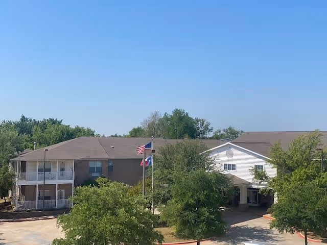 Exterior view of a senior living facility building with a two-story section on the left and a single-story section on the right. There are several trees and a driveway in front of the building. Two flagpoles with the American flag and another flag are visible near the entrance under a clear blue sky.