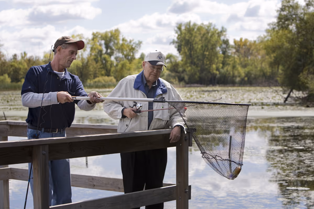 Two men fishing on a wooden dock by a lake surrounded by trees and water lilies. One man is holding a fishing rod while the other is using a large net to catch a fish.