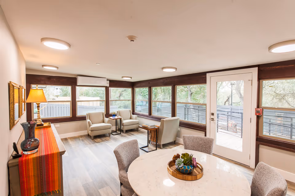 A bright and cozy living room with large windows on two walls, allowing natural light to fill the space. The room features a round white marble table with four upholstered chairs around it, a wooden sideboard with a colorful runner and a lamp, and three beige armchairs arranged near the windows. Outside the windows, trees and a fenced outdoor area are visible.