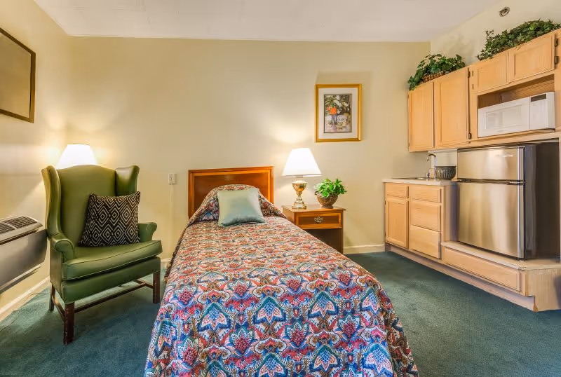A cozy senior living room featuring a single bed with a colorful patterned bedspread, a green armchair with a decorative pillow, a wooden nightstand with a lamp and a potted plant, and a kitchenette area with wooden cabinets, a microwave, and a small refrigerator. The walls are light-colored and there is a framed picture hanging above the nightstand.