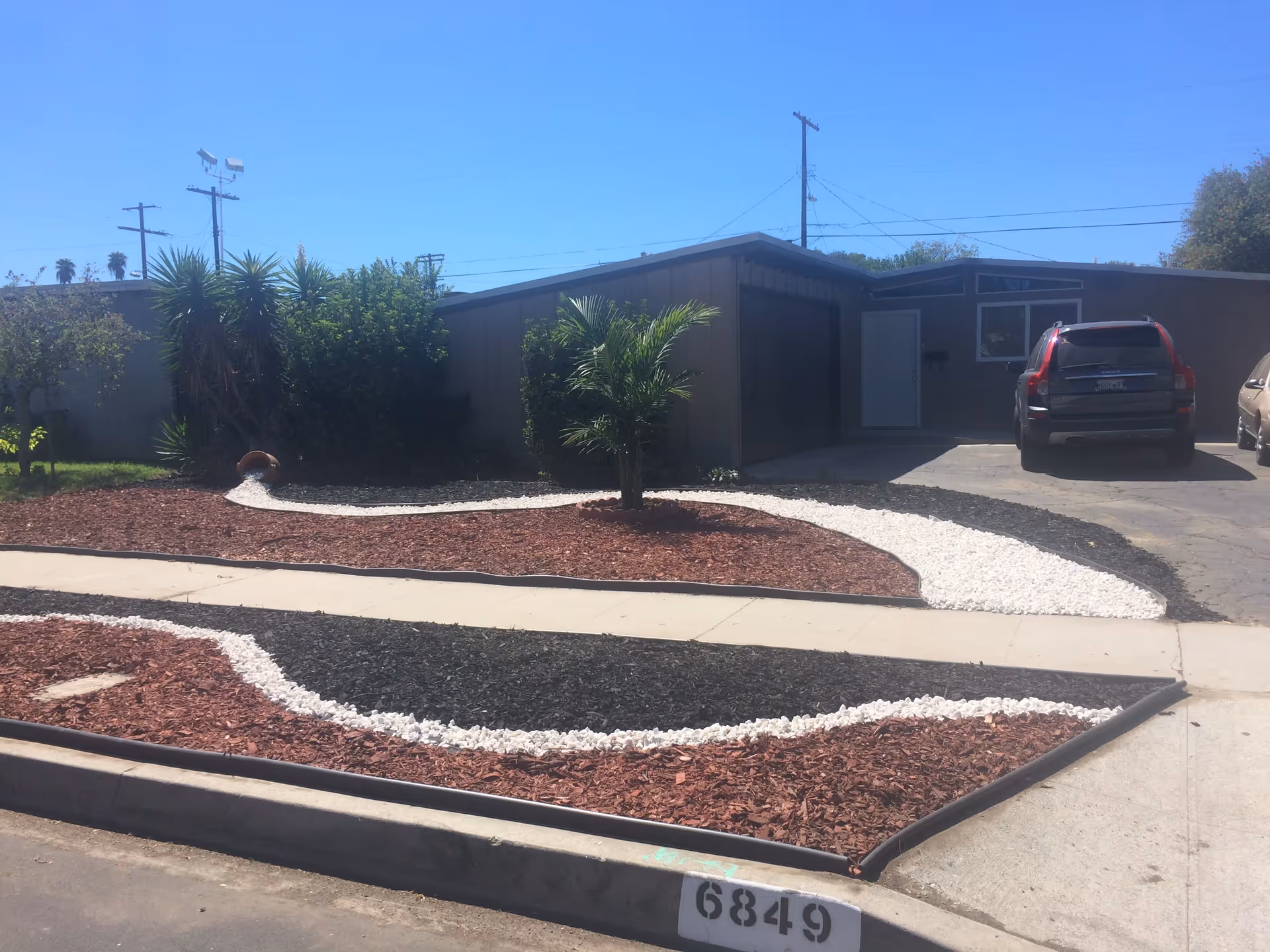 Front yard of a single-story house with a driveway and two parked cars. The yard features decorative landscaping with red and black mulch, white stones arranged in curved patterns, and a small palm tree in the center. The house has a garage with a closed door and a white entry door beside it. The sky is clear and blue.