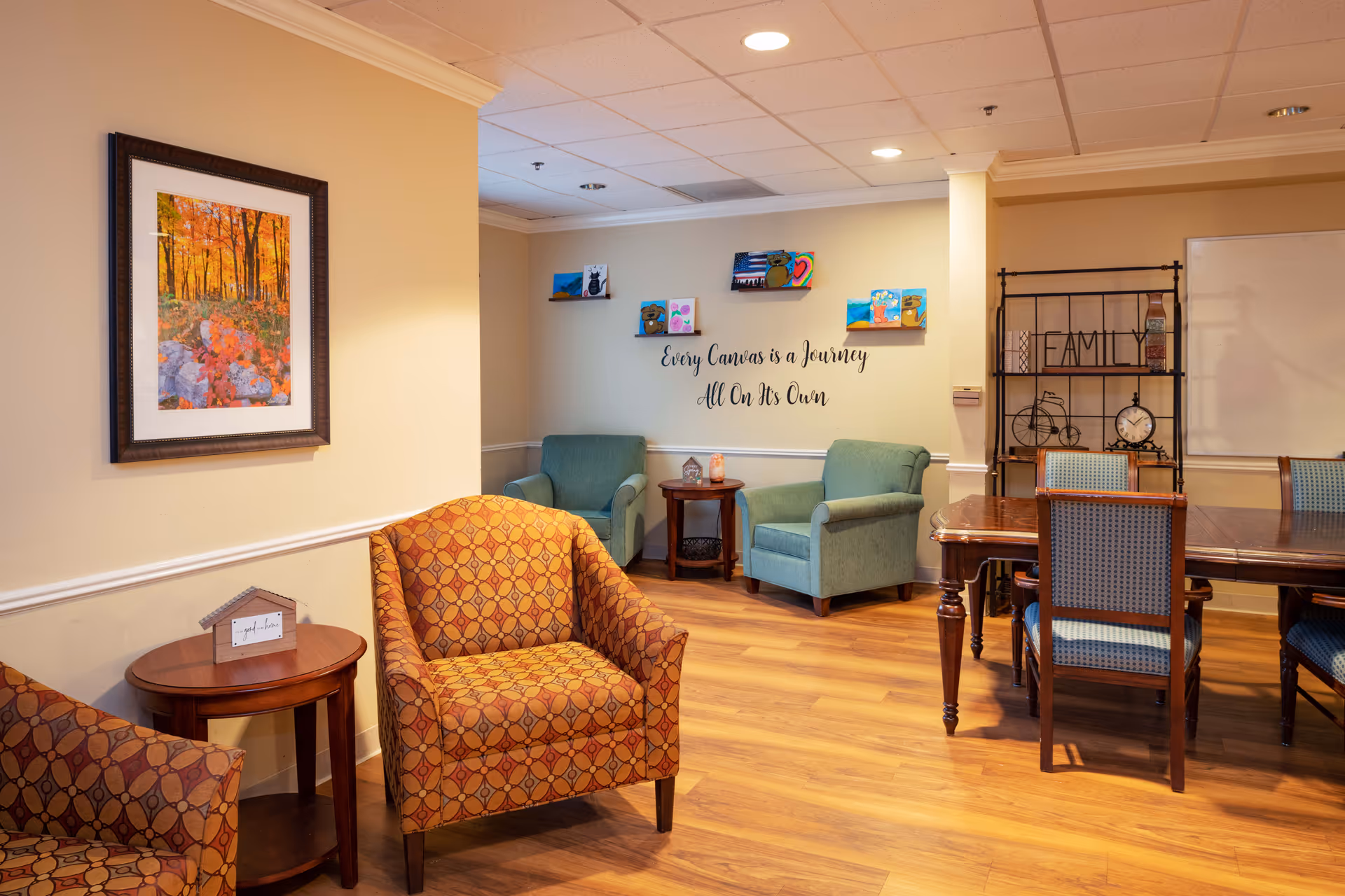 A cozy senior living common area with patterned orange armchairs, two green armchairs, wooden tables, and a wooden dining table with chairs. The walls are decorated with a framed autumn forest photograph, small colorful paintings, and a wall decal that reads 'Every Canvas is a Journey All On Its Own.' There is also a decorative shelving unit with the word 'FAMILY' and various ornaments.