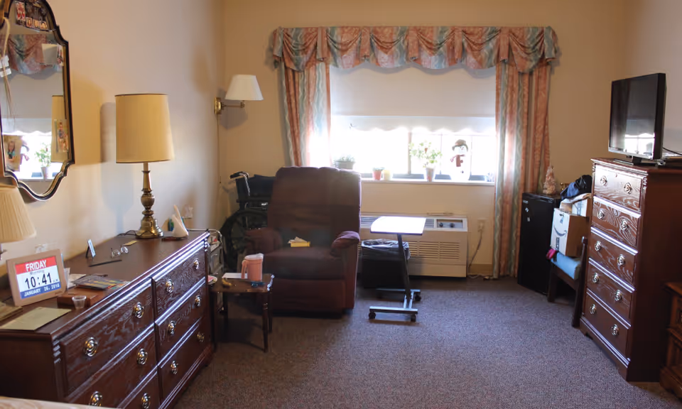 A cozy living room in a senior living facility with a large window covered by patterned curtains. The room contains a brown recliner chair, a small side table with a pink cup, a wooden dresser with a lamp and a digital calendar showing Friday, January 26, 2018, a wheelchair, a TV on a tall wooden chest of drawers, and various small plants on the windowsill.