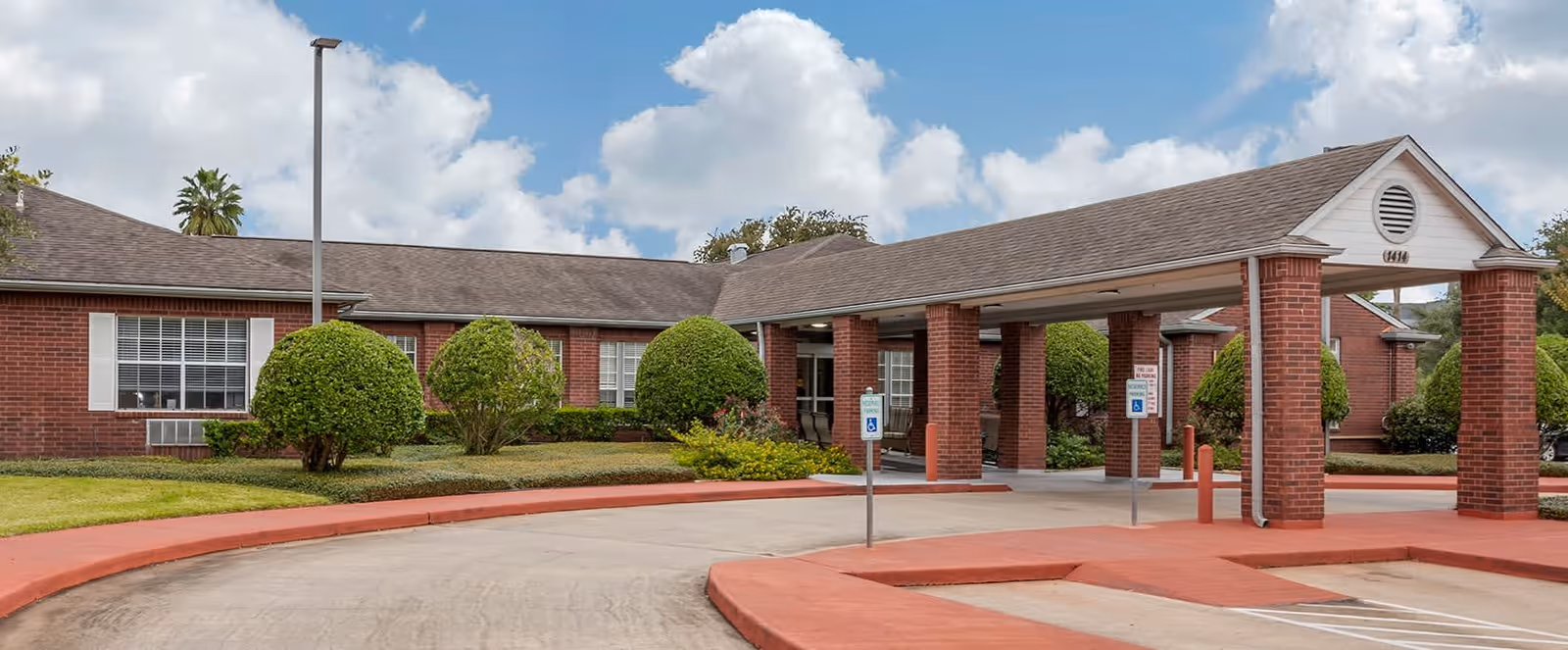 Exterior view of a single-story brick building with a covered entrance supported by brick columns. The building is surrounded by neatly trimmed bushes and a well-maintained lawn under a partly cloudy sky.