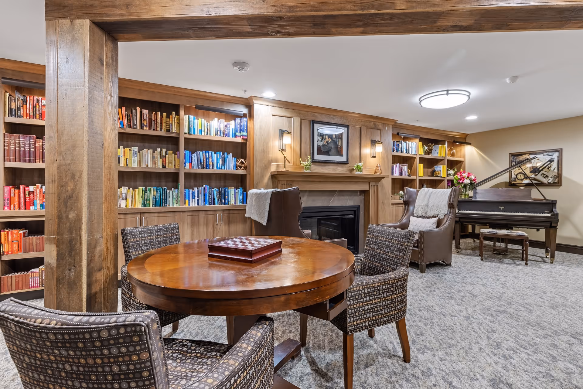 A cozy living room area with a round wooden table and four patterned chairs in the foreground. Behind the table, there is a fireplace with a wooden mantel, flanked by built-in bookshelves filled with books arranged by color. Two leather armchairs with blankets draped over them are positioned near the fireplace. In the background, there is a grand piano with a bench and a framed artwork on the wall. The room has carpeted flooring and warm lighting from ceiling fixtures and wall sconces.