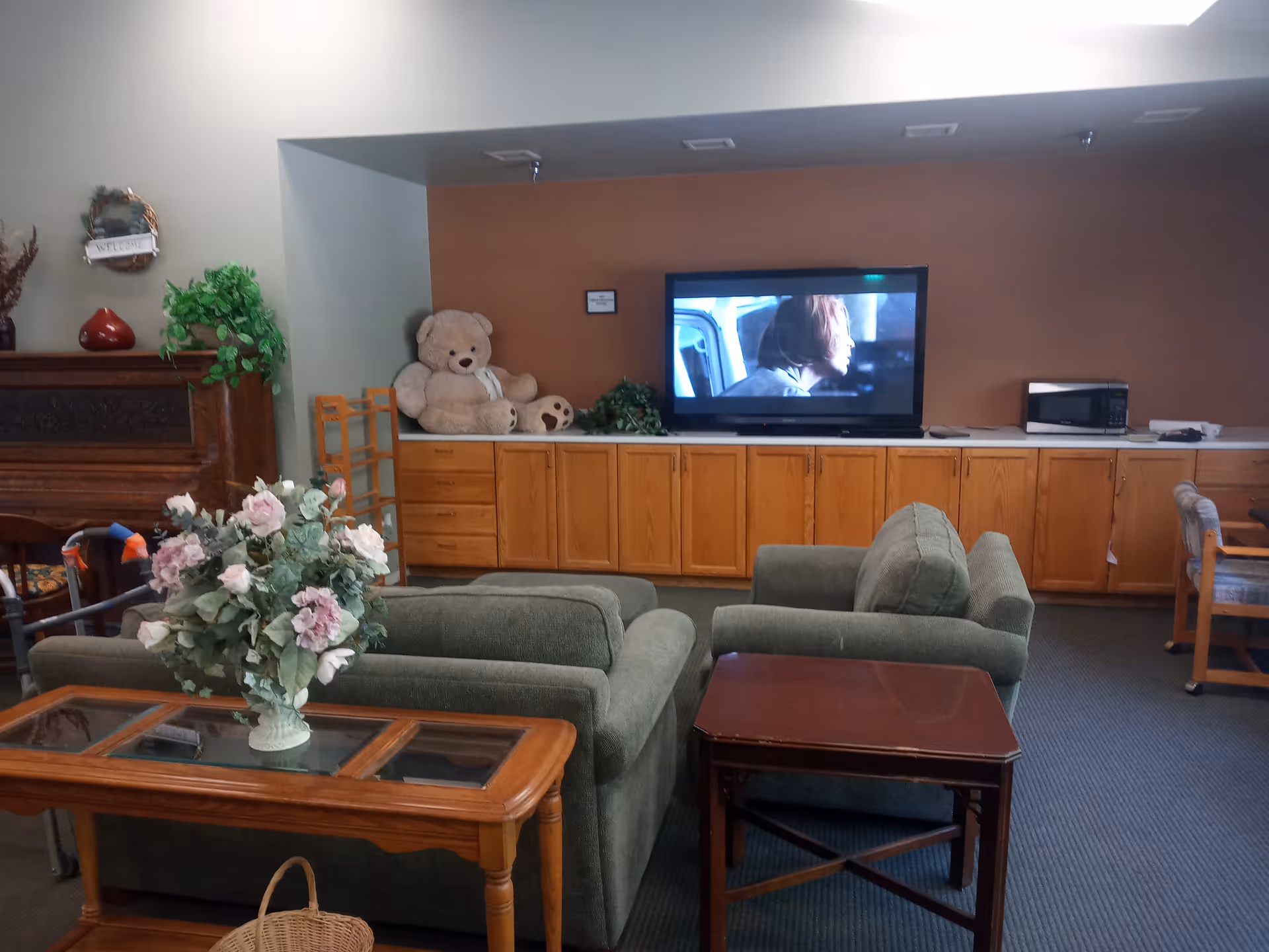A cozy living room area in a senior living facility with two green upholstered armchairs facing a flat-screen TV mounted on a brown wall. Below the TV is a long wooden cabinet with multiple drawers and a microwave on the right side. A large teddy bear and some plants are placed on the cabinet. In the foreground, there is a wooden coffee table with a glass top and a vase of artificial flowers. The room has a carpeted floor and a fireplace with decorative items on the left side.