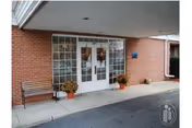 Entrance to a building with double glass doors framed by large windows on either side. There is a bench to the left of the doors and two potted plants with flowers placed near the entrance. The building exterior is made of red brick and there is a covered overhang above the entrance.
