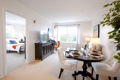 Bright and spacious living area in a senior living facility with a round dining table set for four, a sitting area with a sofa and coffee table, a TV on a cabinet, and a large window letting in natural light. A bedroom with a bed and bedside lamp is visible through an open doorway.
