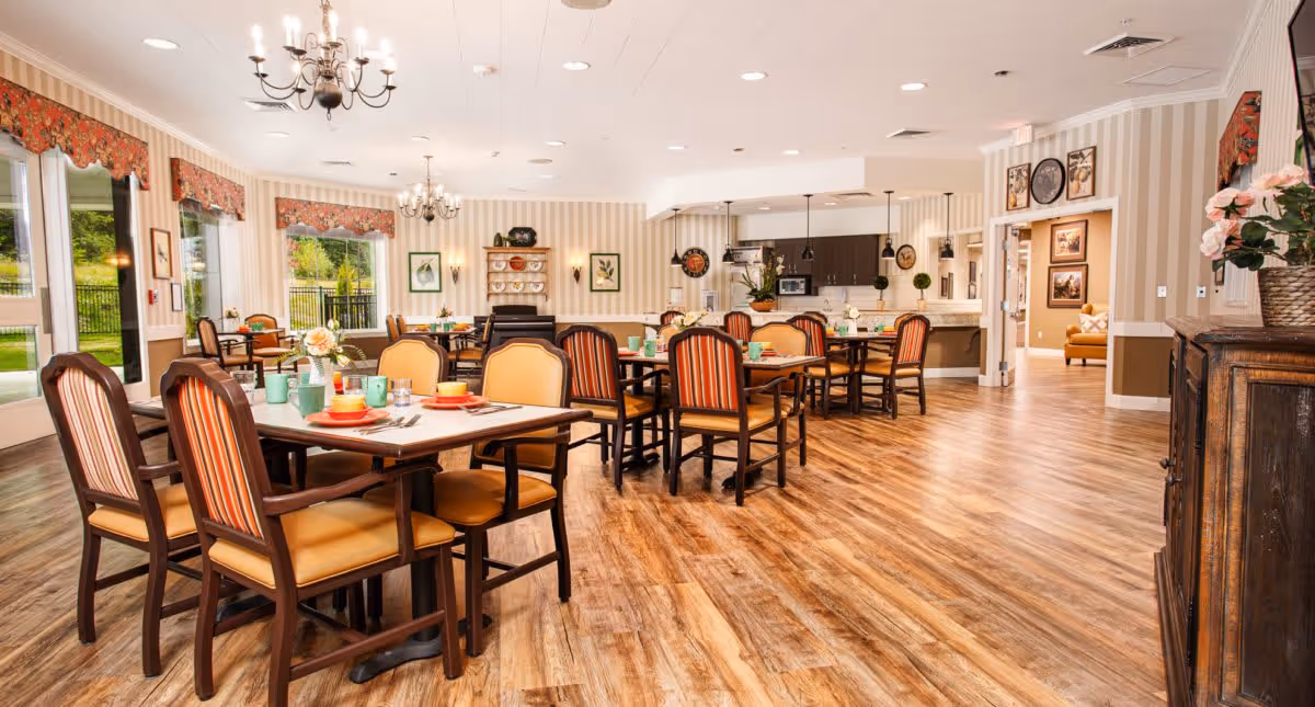 A bright and spacious dining room with wooden floors, multiple tables set with colorful dishes and cups, upholstered chairs with striped and solid patterns, chandeliers hanging from the ceiling, large windows with floral valances letting in natural light, and a kitchen area in the background.