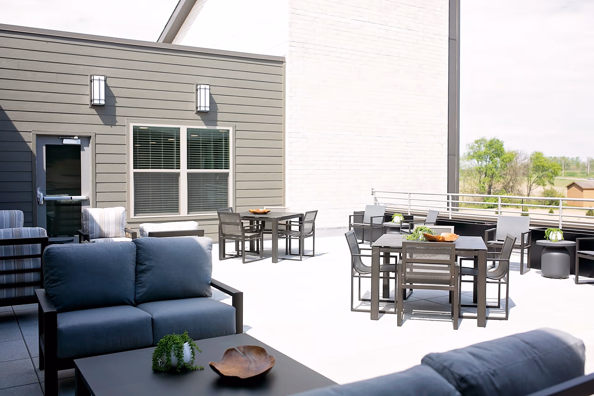 Outdoor patio area with modern seating including cushioned sofas, chairs, and tables. The space is bright and open with a railing overlooking greenery in the distance. There are decorative plants on the tables and the building exterior is visible with a door and windows.