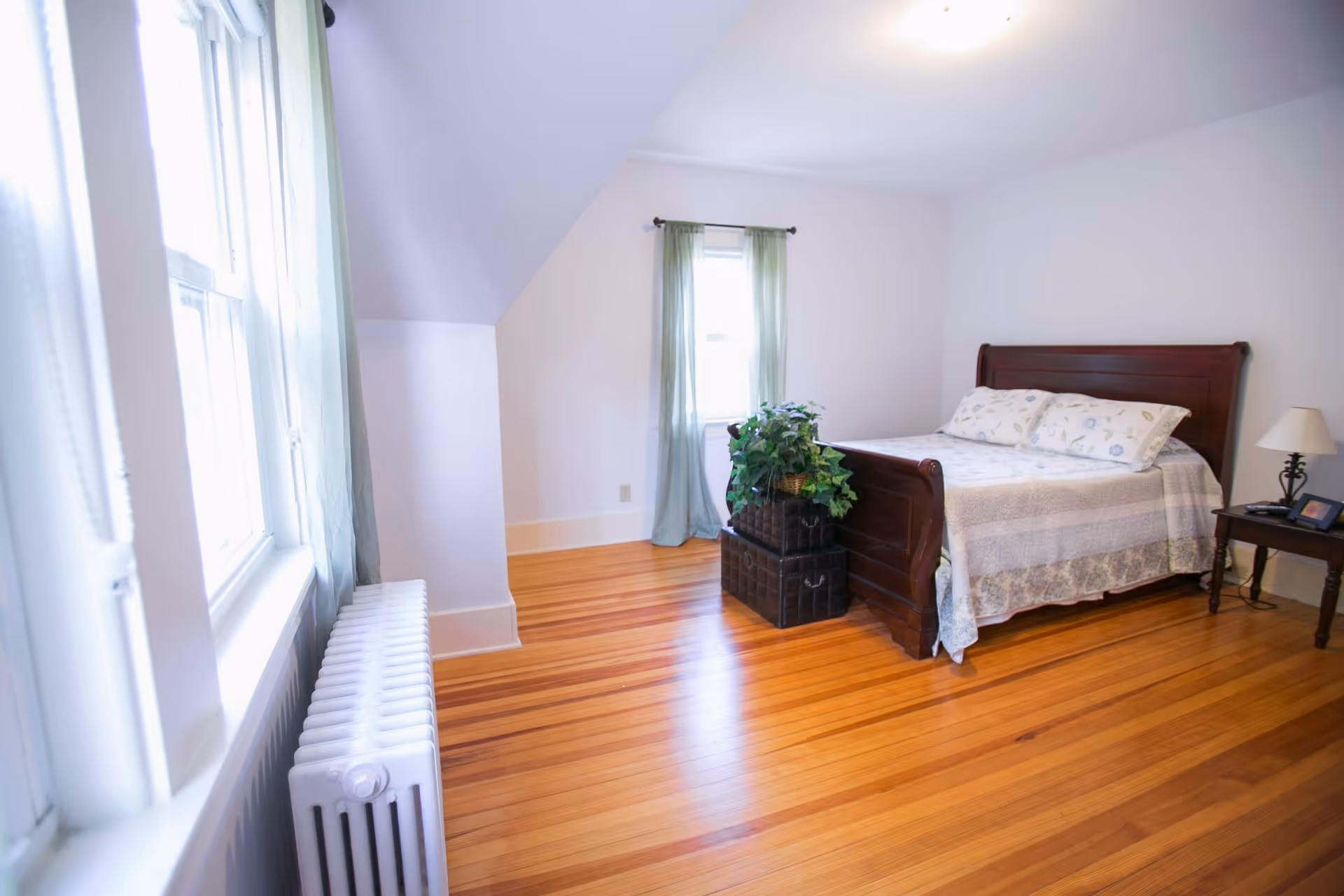 A bright bedroom with wooden floors, a wooden bed with white and patterned bedding, a small wooden side table with a lamp and picture frame, a window with light green curtains, and a white radiator beneath another window.