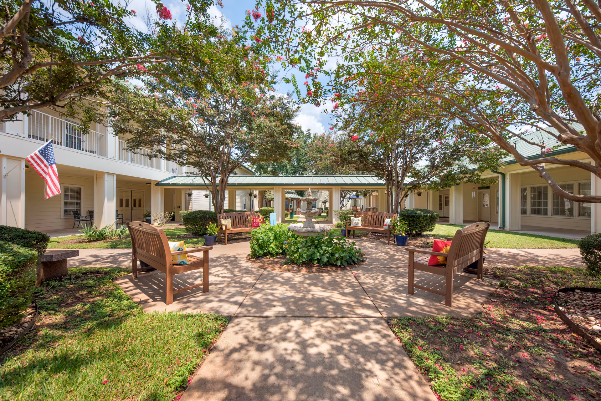 Outdoor courtyard area at Brookdale Beckett Meadows featuring wooden benches with colorful cushions arranged around a central fountain. The space is shaded by trees with pink flowers and surrounded by a two-story building with balconies and an American flag displayed.