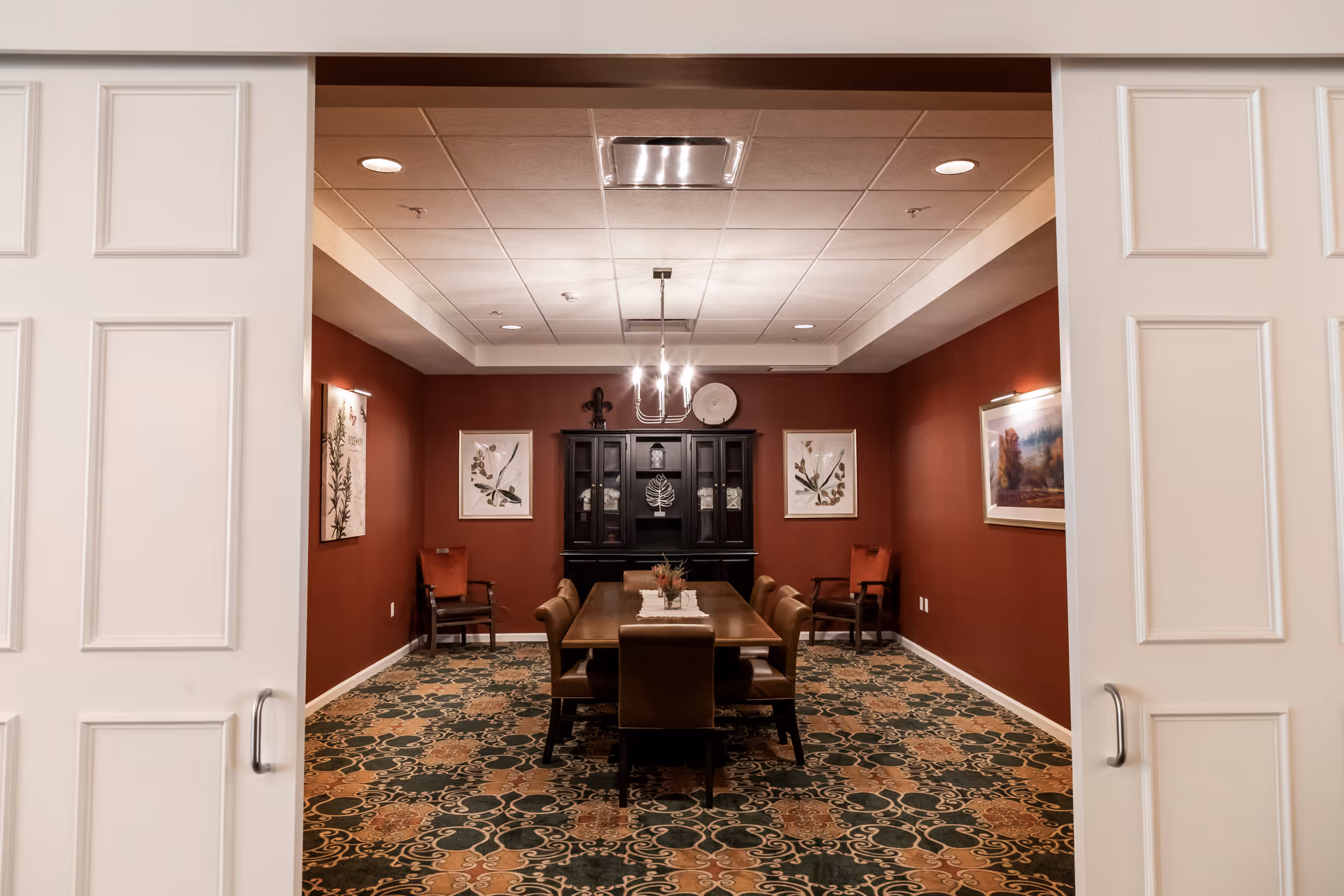 A formal dining room with a long wooden table surrounded by eight chairs. The walls are painted a deep red and decorated with framed artwork. At the far end of the room is a dark wooden cabinet with decorative items on top. The room is lit by a modern chandelier hanging from a white ceiling with recessed lighting. The floor is covered with a patterned carpet in green, beige, and brown tones. The room is viewed through partially open white double doors.