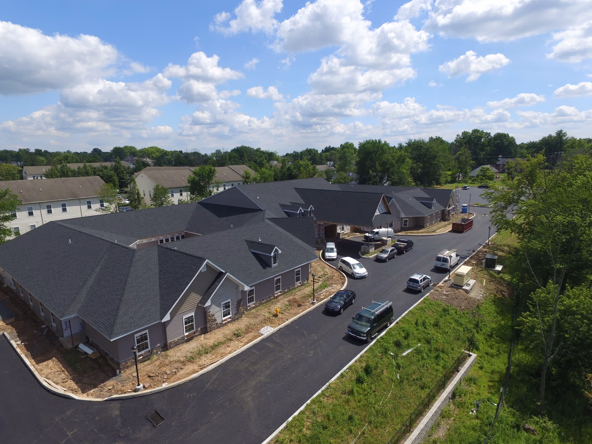 Aerial view of a large single-story building with dark gray roofs and beige siding, surrounded by a newly paved driveway with several parked vehicles. The building is situated in a green, suburban area with trees and other residential buildings in the background under a partly cloudy sky.