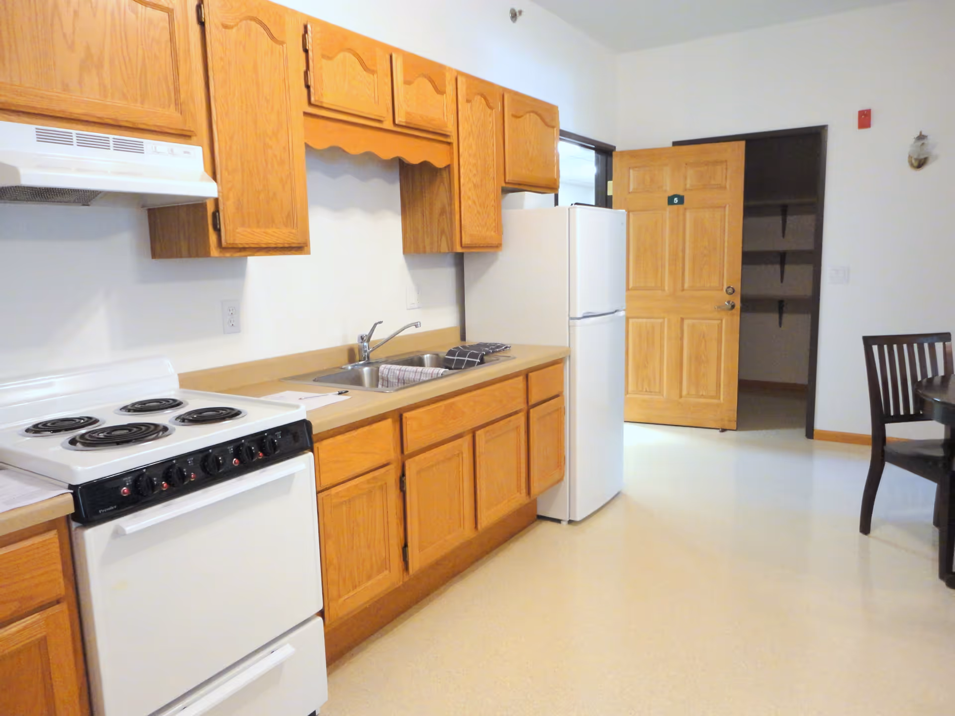 A kitchen area with wooden cabinets, a white electric stove, a double sink with a dish towel, and a white refrigerator. There is an open wooden door leading to a pantry or storage area with shelves. Part of a dining table and chair are visible on the right side.