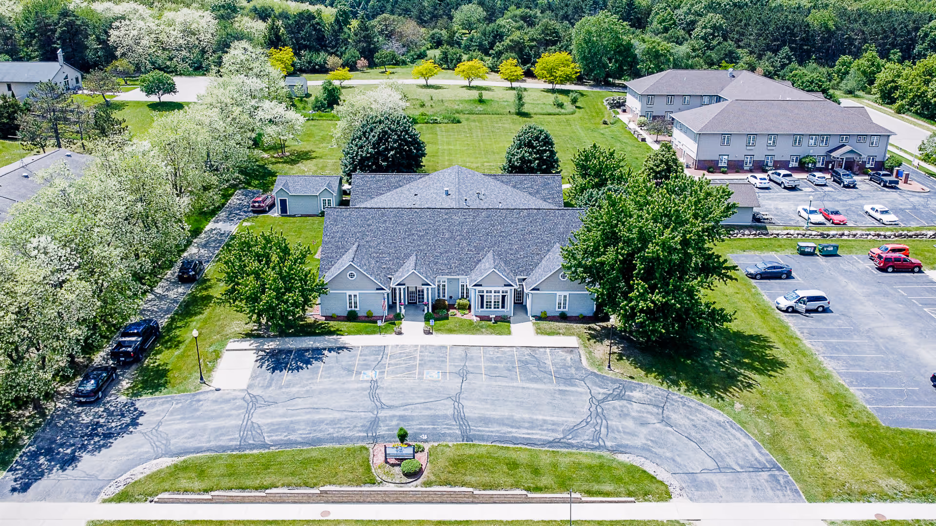 Aerial view of a senior living facility showing a large building with a gray roof surrounded by green lawns and trees. There is a parking lot in front of the building with several cars parked. Additional buildings and parking areas are visible in the background.