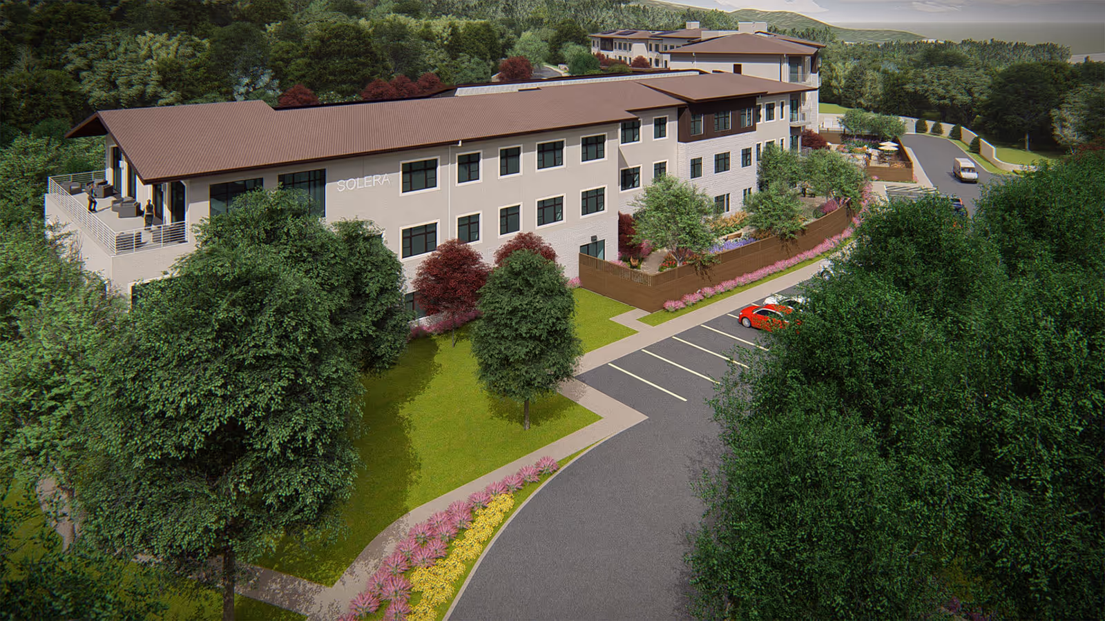Aerial view of a senior living facility named Solera, showing a three-story building surrounded by trees and landscaped gardens. There is a parking lot with a few cars and a driveway leading to the building. The building has a brown roof and beige exterior walls with multiple windows. People are visible on a balcony on the left side of the building.