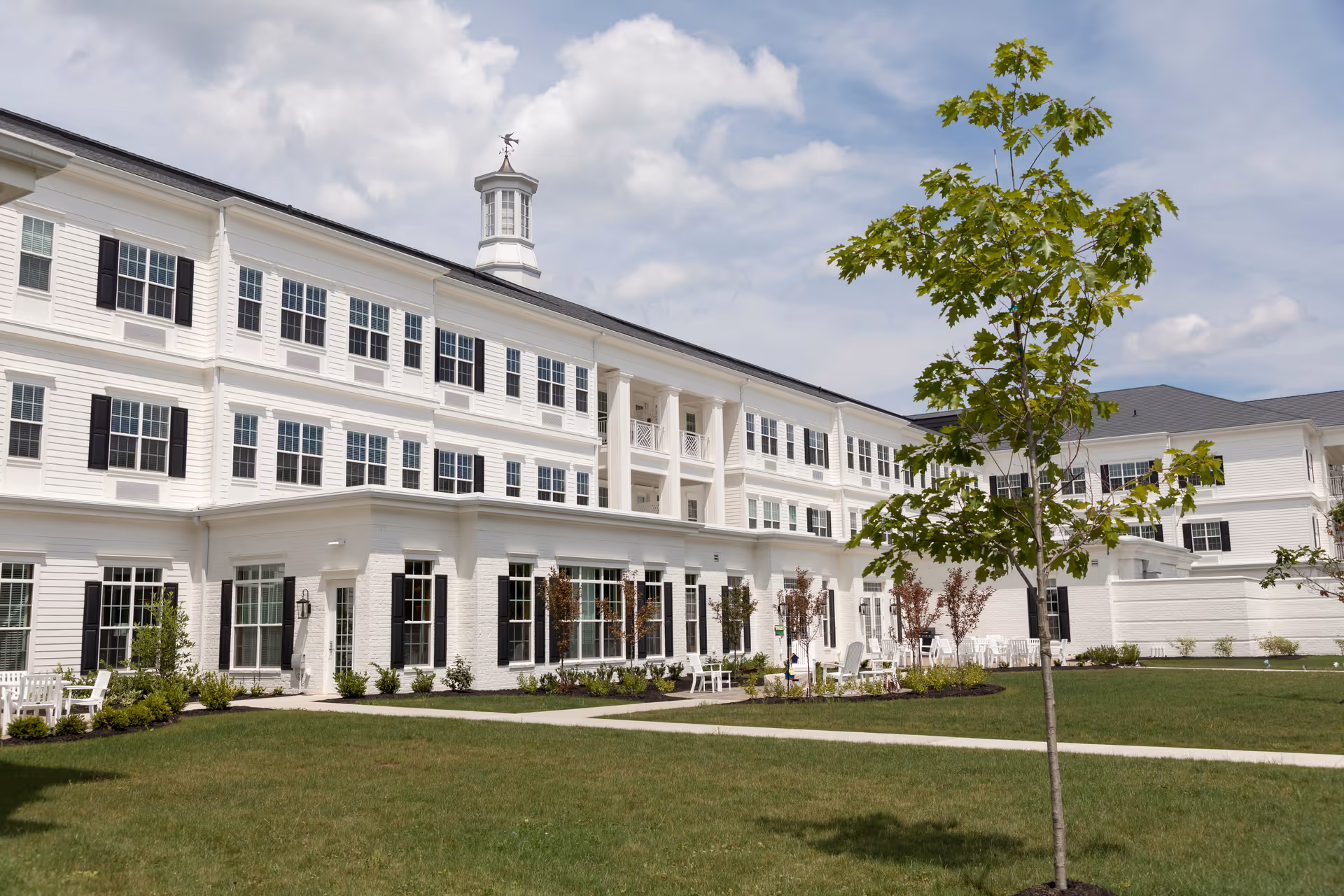 Exterior view of a large white multi-story senior living facility building with many windows and black shutters. The building has a cupola on the roof and is surrounded by a well-maintained lawn with a young tree and outdoor seating areas. The sky is partly cloudy.