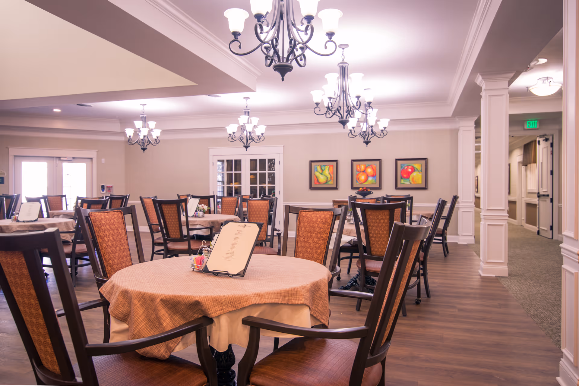 A well-lit dining room in a senior living facility with multiple round tables covered with beige tablecloths and surrounded by wooden chairs with patterned upholstery. The room features elegant chandeliers hanging from the ceiling, framed colorful fruit paintings on the wall, and a carpeted hallway leading to other areas.
