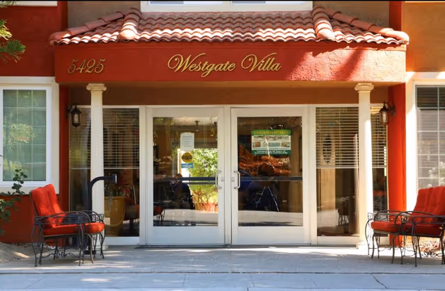 Entrance of Westgate Villa Assisted Living facility with double glass doors, red cushioned chairs on either side, and a red tiled roof above the doorway. The building number 5425 is displayed on the left side of the entrance.