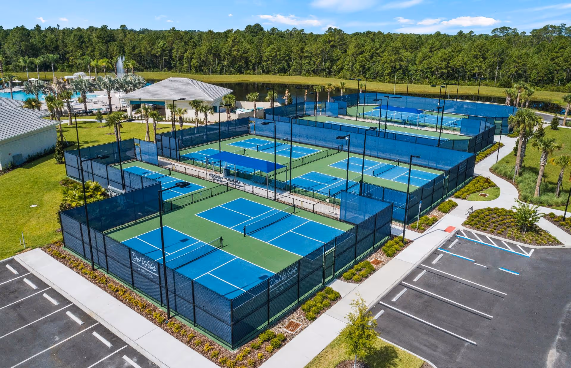 Aerial view of multiple fenced pickleball courts with blue and green surfaces at Del Webb Sunbridge retirement community, surrounded by landscaped greenery, parking spaces, and a small pavilion near a pond with trees in the background.