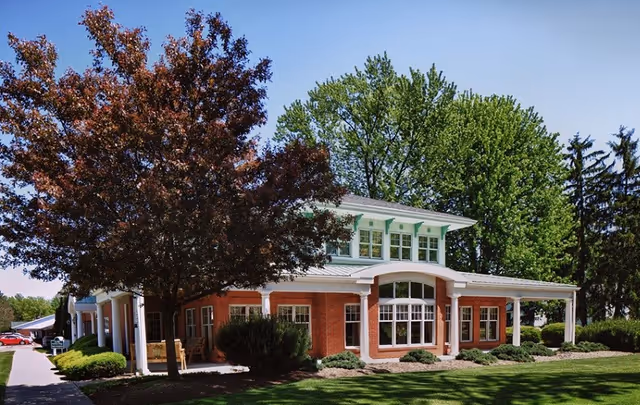 A two-story brick building with white trim and large windows surrounded by green trees and shrubs under a clear blue sky, with a sidewalk and a red car visible on the left side.